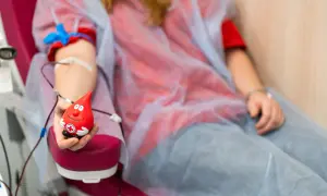 Uśmiechnięta kropla krwi trzymająca zabawkę, symbol tego, czy oddawanie krwi jest zdrowe. Osoba w czerwonej koszulce i niebieskim fartuchu oddaje krew.