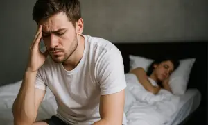 Mężczyzna zmartwiony, siedzi na łóżku. Obok śpi kobieta. Może to być problem z brakiem wzwodu po testosteronie.