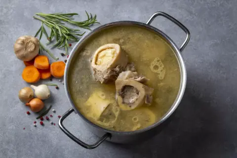 Garnek z bulionem, w którym pływają kości szpikowe na rosół. Obok marchewka, czosnek, cebula i rozmaryn.