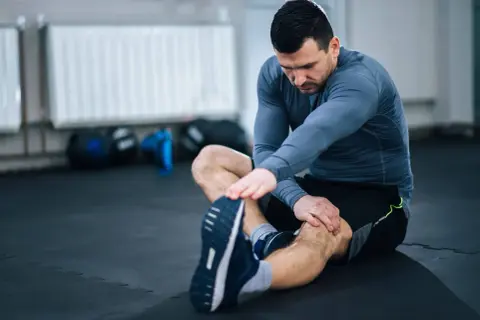 Mężczyzna wykonuje ćwiczenia rozciągające nogi na siłowni, siedząc na macie.